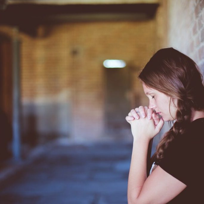 woman praying while leaning against brick wall